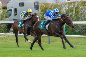 Two jockeys race neck-and-neck on galloping horses; lead horse wears green number 1 saddle cloth, rider in blue. Behind, a horse with green number 2 and a yellow-silk rider follows.