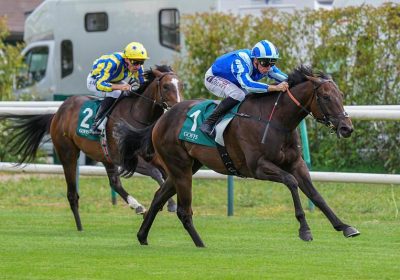 Two jockeys race neck-and-neck on galloping horses; lead horse wears green number 1 saddle cloth, rider in blue. Behind, a horse with green number 2 and a yellow-silk rider follows.